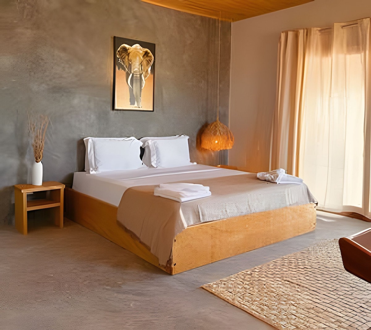 A spacious sleeping area features a large wooden bed dressed in white linens and a neutral-colored blanket. Natural light filters through sheer curtains, illuminating the room. An artistic elephant print adorns the wall above the bed, complemented by a woven pendant light and a simple nightstand.