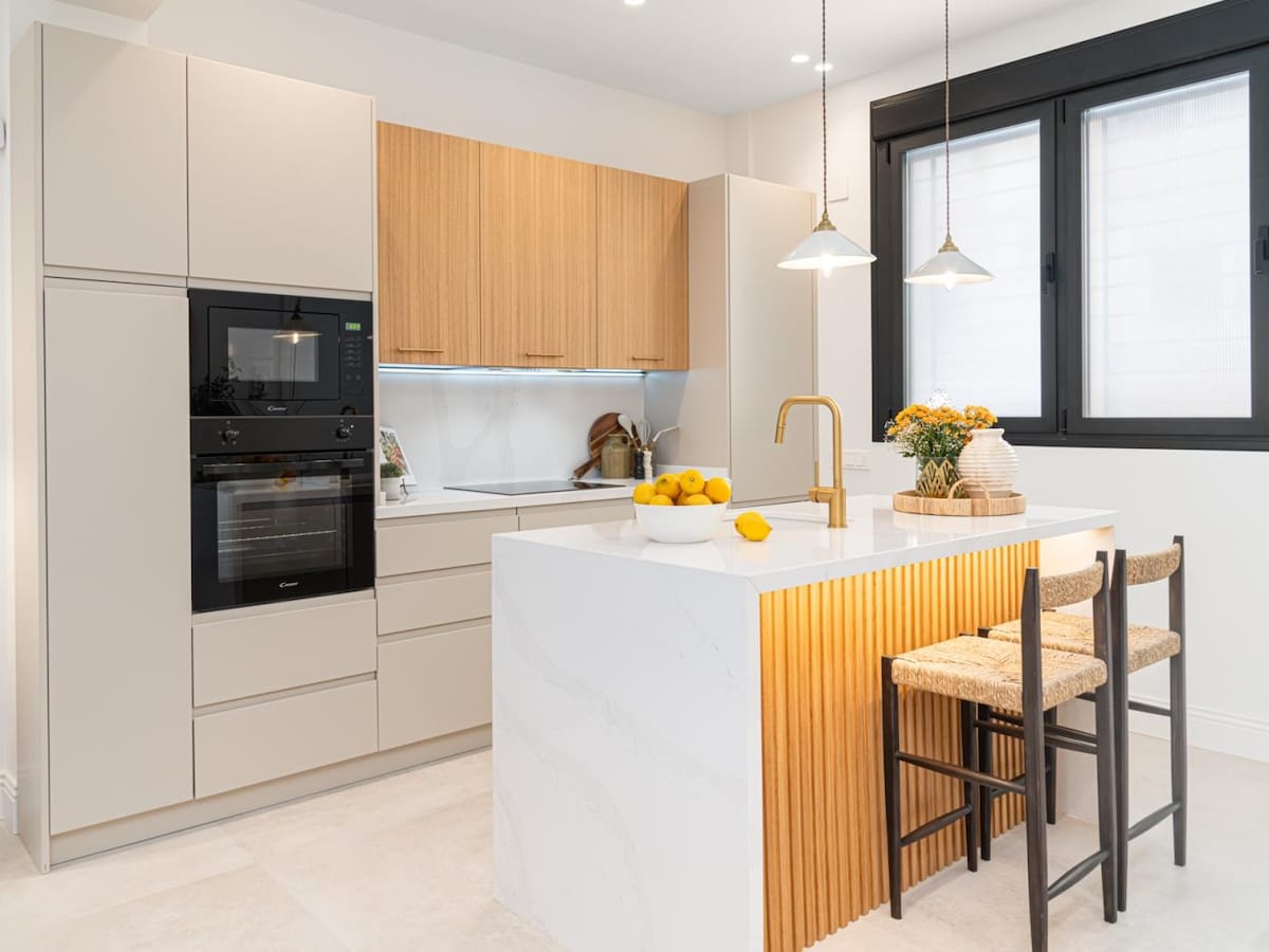 A modern kitchen equipped with sleek cabinetry and a spacious island featuring a textured wooden front. Appliances include an oven and microwave built into the cabinetry. Two high stools are positioned at the island, alongside a decorative arrangement of oranges and a small vase of flowers.