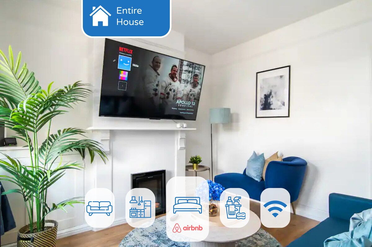 A modern living room is presented with a large wall-mounted flat-screen TV featuring streaming options. A cozy seating arrangement is highlighted by blue accents and a green plant, with natural light illuminating the space. Decorative artwork enhances the wall above a fireplace.
