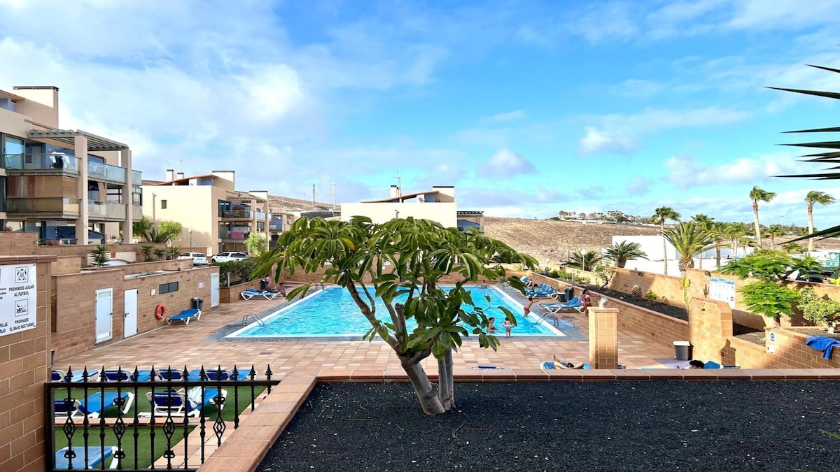 A spacious outdoor area features a large swimming pool framed by lounge chairs. Surrounding palm trees and greenery add a natural element, while a sunlit sky enhances the bright ambiance of the space. The pool area is designed for relaxation and leisure.