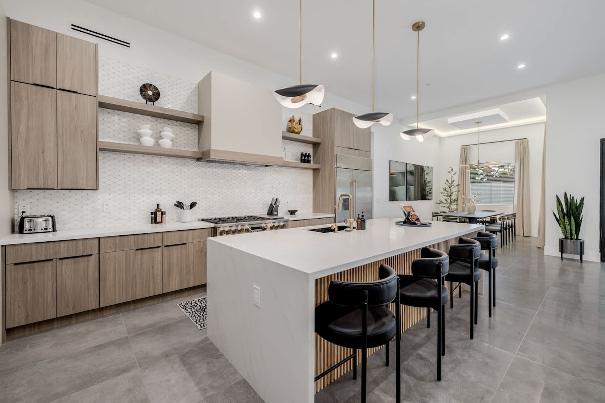 A modern kitchen features a large island with seating for six, accented by elegant pendant lighting. Customized cabinetry offers ample storage, while high-end appliances and a stylish backsplash enhance the space. Neutral tones and sleek surfaces contribute to a sophisticated ambiance.