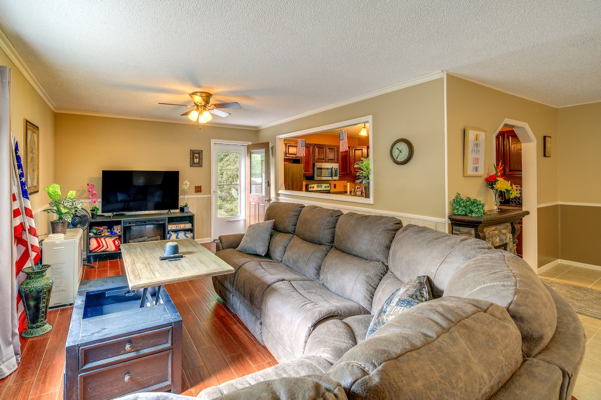 A spacious living area features a large sectional sofa arranged around a wooden coffee table. A flat-screen TV is positioned on an adjacent wall, and natural light enters through a doorway leading outside. Decorative accents enhance the welcoming atmosphere.