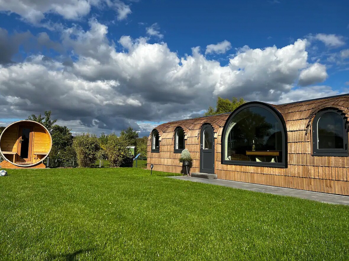 A uniquely designed lodge features a thatched exterior and large arched windows, surrounded by a lush green lawn. A sauna with a circular structure stands nearby, framed by a backdrop of partly cloudy skies.