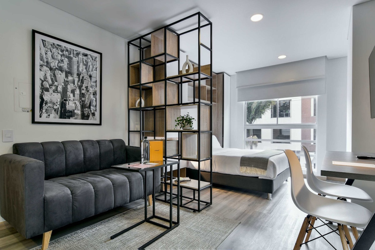 A modern studio highlights a comfortable sofa accompanied by a small table and sleek chairs. Natural light fills the space, enhanced by the open layout. A stylish bookshelf separates the living area from the sleeping area, showcasing organized decor and greenery, while large windows overlook the surroundings.