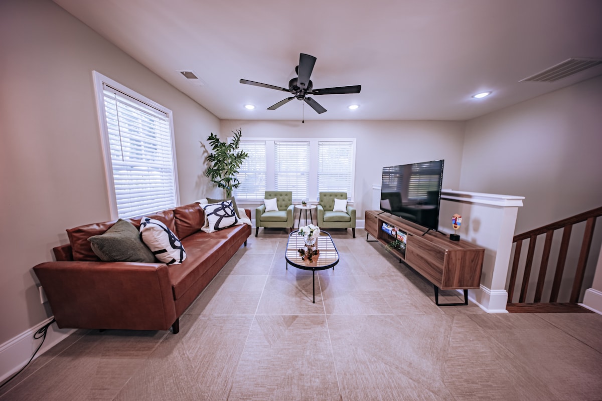 An open-concept living area is highlighted by a spacious leather sofa and stylish green armchairs, creating a cozy setting. A large flat-screen TV is mounted on the wall, and a modern coffee table occupies the center, while natural light filters through the window blinds.