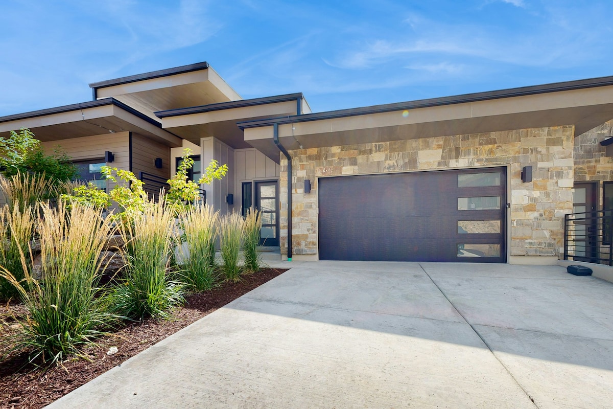The exterior of a modern house is depicted, featuring a combination of stone and wood elements. A spacious driveway leads to a stylish garage door. Lush grasses and carefully curated landscaping create an inviting entrance, complemented by clear blue skies above.