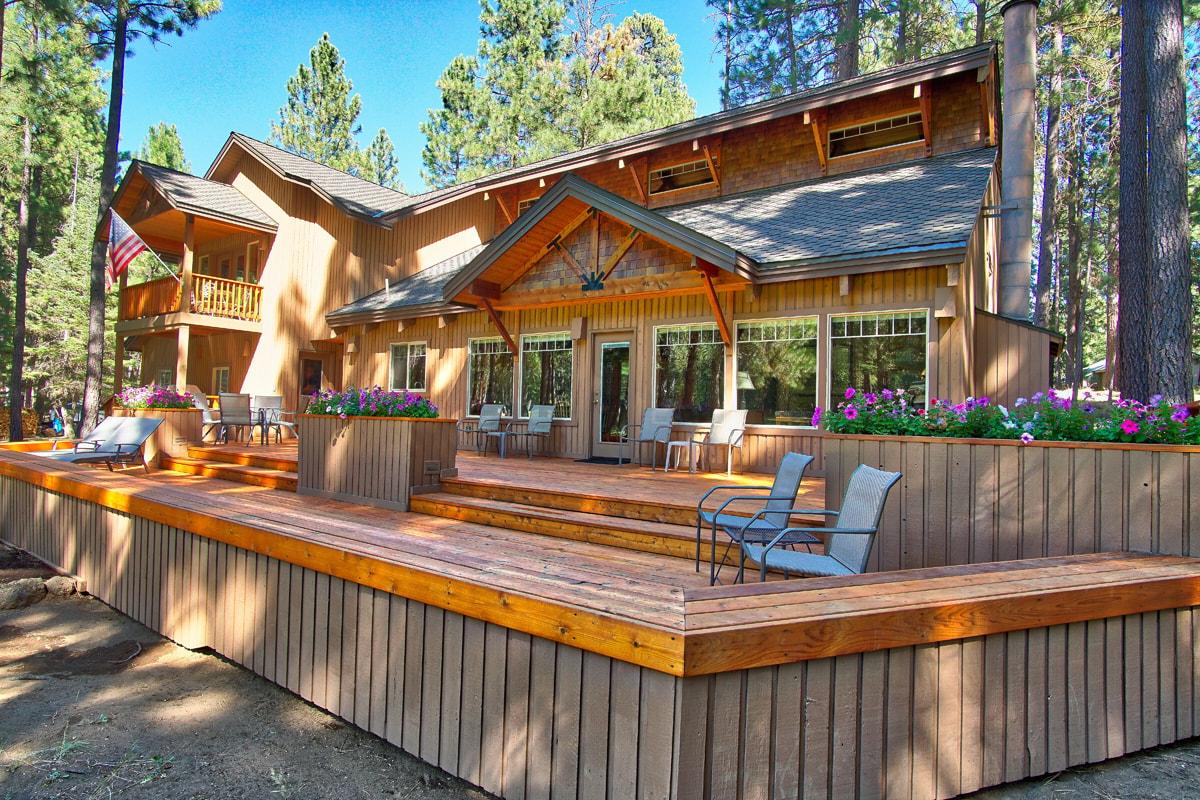 The exterior of the home features a spacious deck with multiple seating options, surrounded by vibrant flowers. The warm wood tones of the façade complement the natural setting of tall trees, providing a serene and inviting atmosphere.