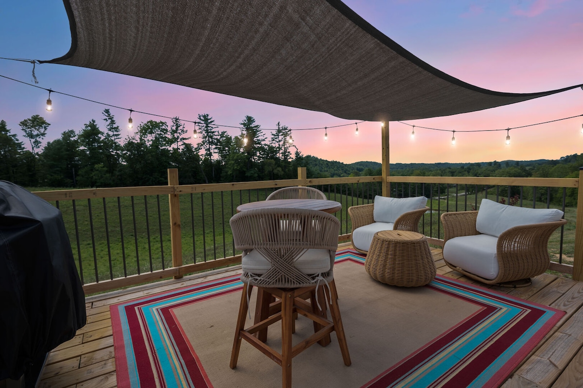 A spacious deck is highlighted by comfortable seating arrangements and a dining set. String lights hang above, creating a relaxed atmosphere. The view reveals expansive greenery under a colorful sky during twilight, inviting moments of relaxation and connection.