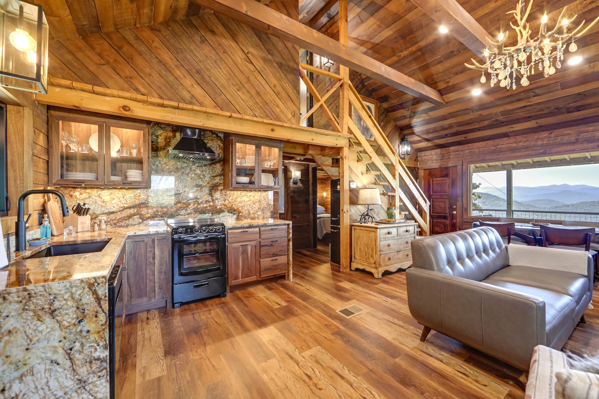 A spacious kitchen and living area are showcased with wooden cabinetry and a marble countertop. A gas stove is prominently displayed, with a comfortable couch and a chandelier adding to the cozy atmosphere. Large windows provide views of the surrounding mountains.