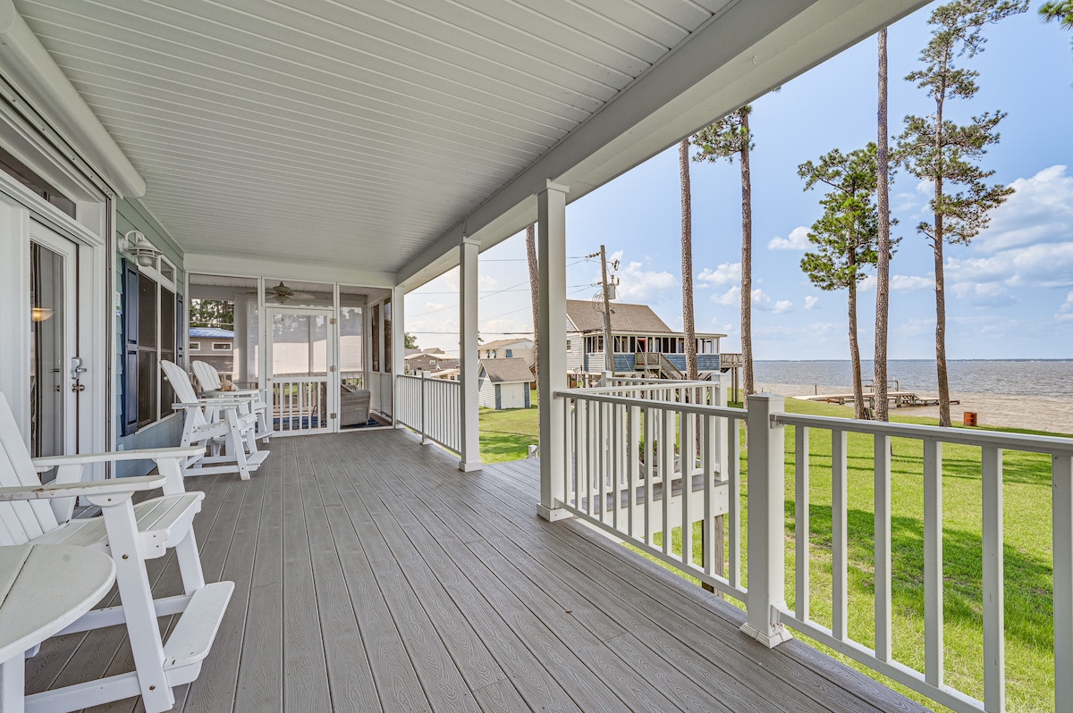 A spacious screened porch is showcased, featuring white rocking chairs positioned to take in views of the Pamlico River. Tall trees provide shade, while the expansive lawn leads towards the water, creating an inviting outdoor space for relaxation.