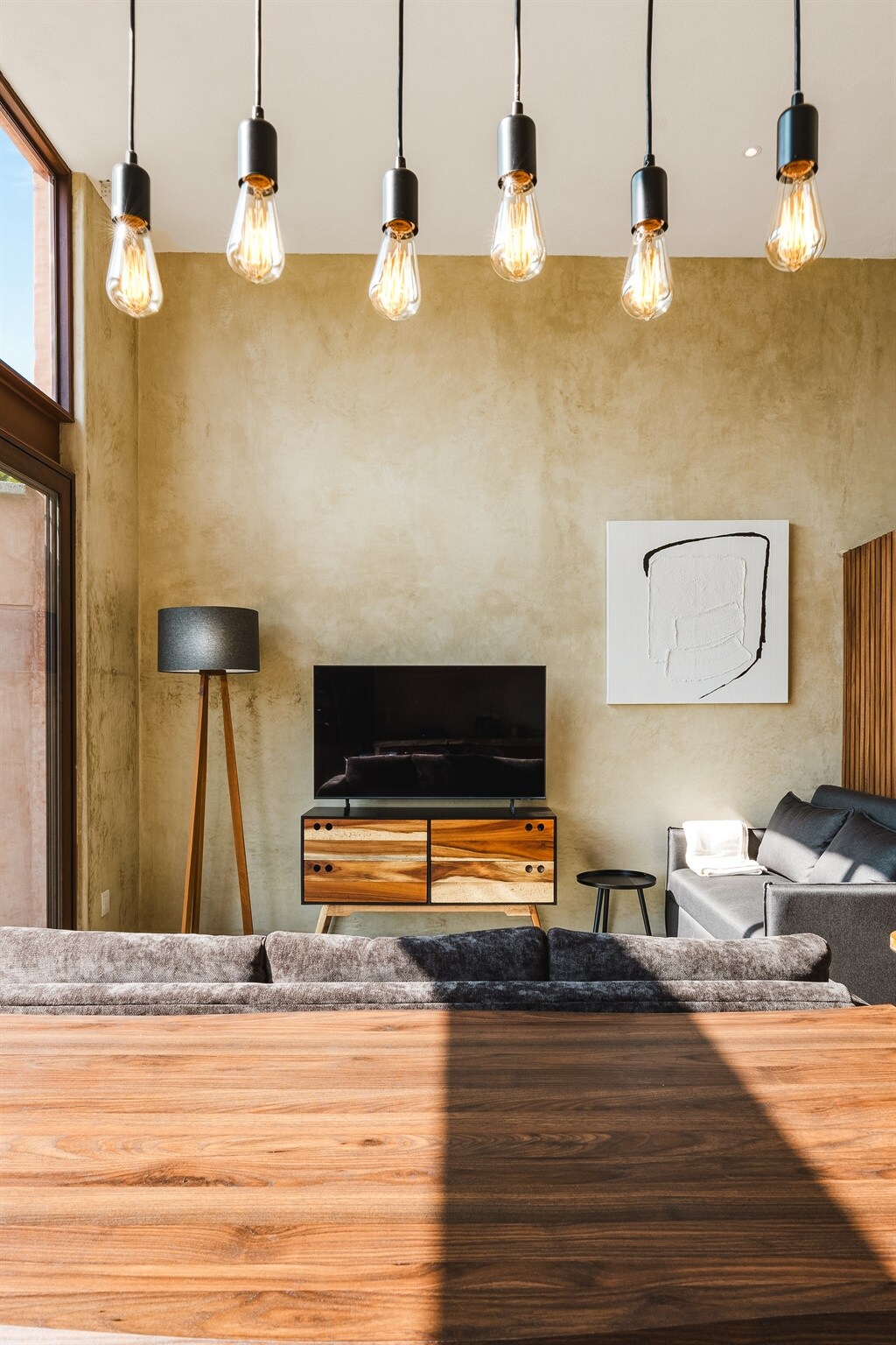 A stylish living area features a comfortable dark sofa positioned near a modern television stand. Above, pendant lights illuminate the space, and a tall floor lamp adds additional lighting. A wall adorned with artwork complements the neutral tones of the interior.