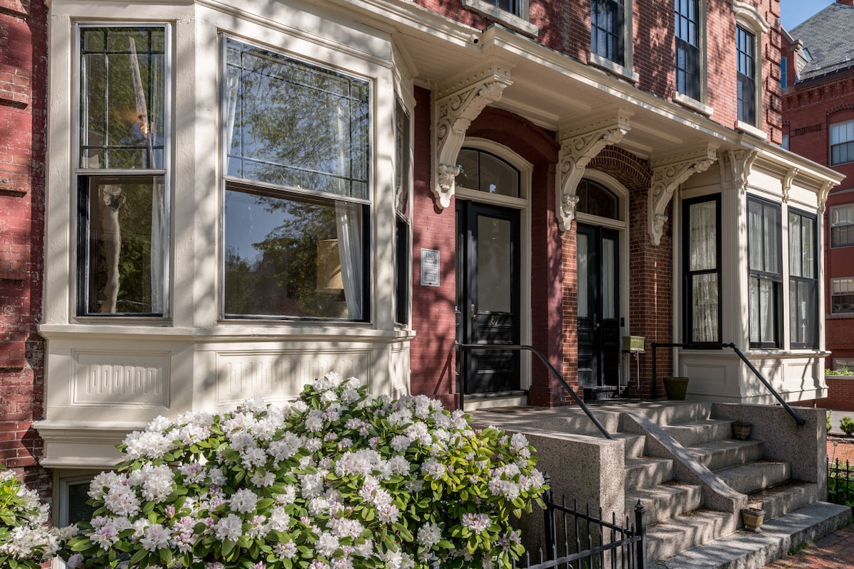 The historic entrance features a grand façade with ornamental detailing and large bay windows. Lush greenery includes blooming flowers in the foreground, accompanied by a stone staircase leading to the front doors. The architecture exhibits traditional craftsmanship and a welcoming atmosphere.