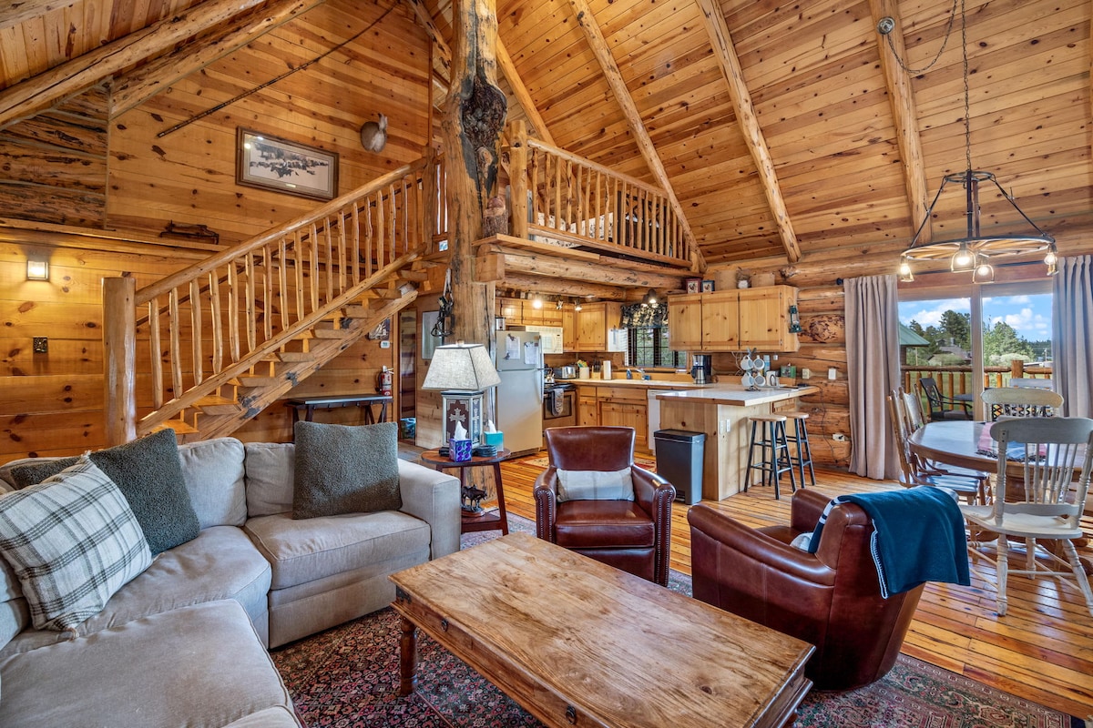 An airy living area showcases a spacious sofa and two armchairs surrounding a wooden coffee table. Vaulted ceilings with wooden beams draw attention to the upper-level loft. Light enters through large windows, illuminating the rustic decor and inviting atmosphere.