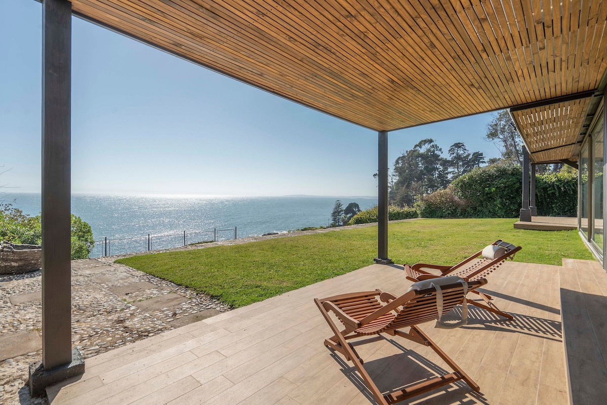 A spacious outdoor area features a wooden deck with lounge chairs positioned to overlook the ocean. Sunlight reflects on the water, creating a inviting seaside view. Lush greenery surrounds the space, enhancing the connection with nature.