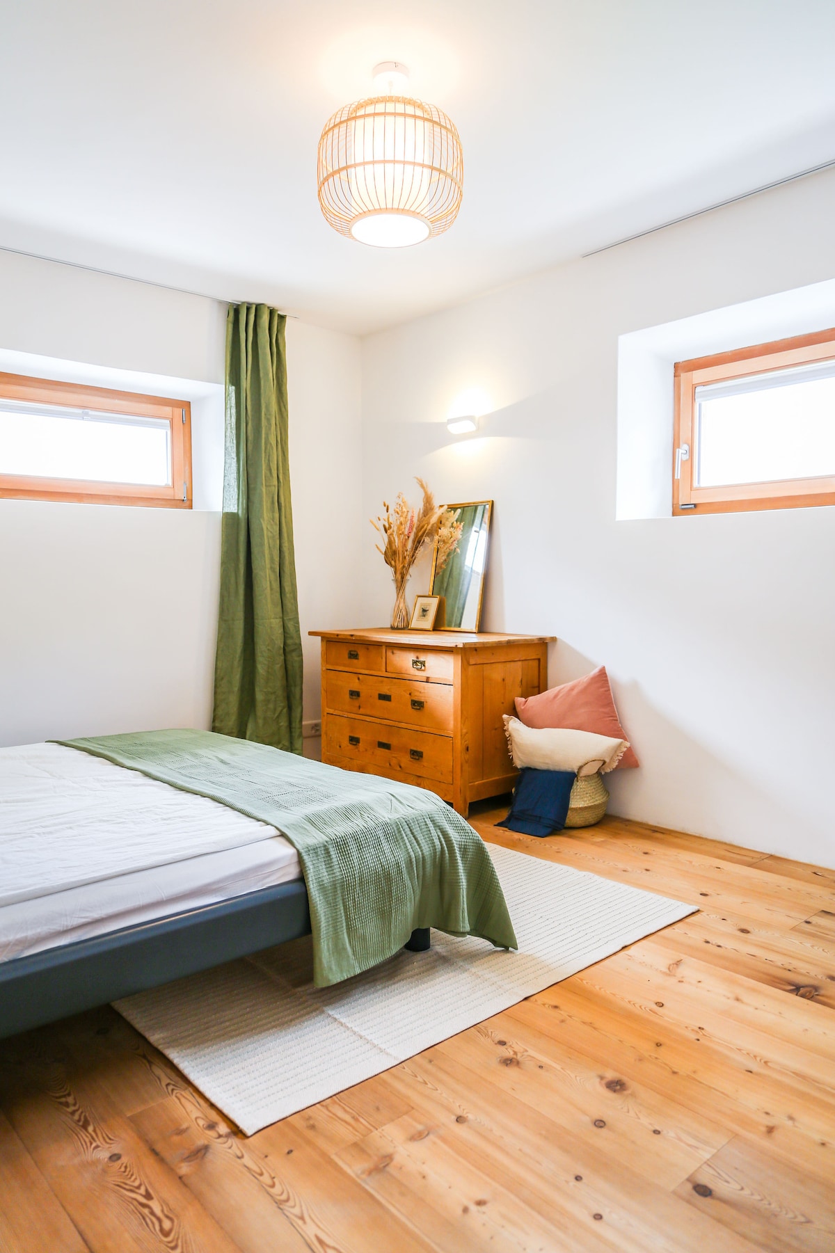 A serene bedroom features a spacious bed with neutral bedding. Natural light enters through two small windows, highlighting the wooden flooring. A vintage dresser stands against the wall, complemented by a decorative mirror and a cozy arrangement of cushions.