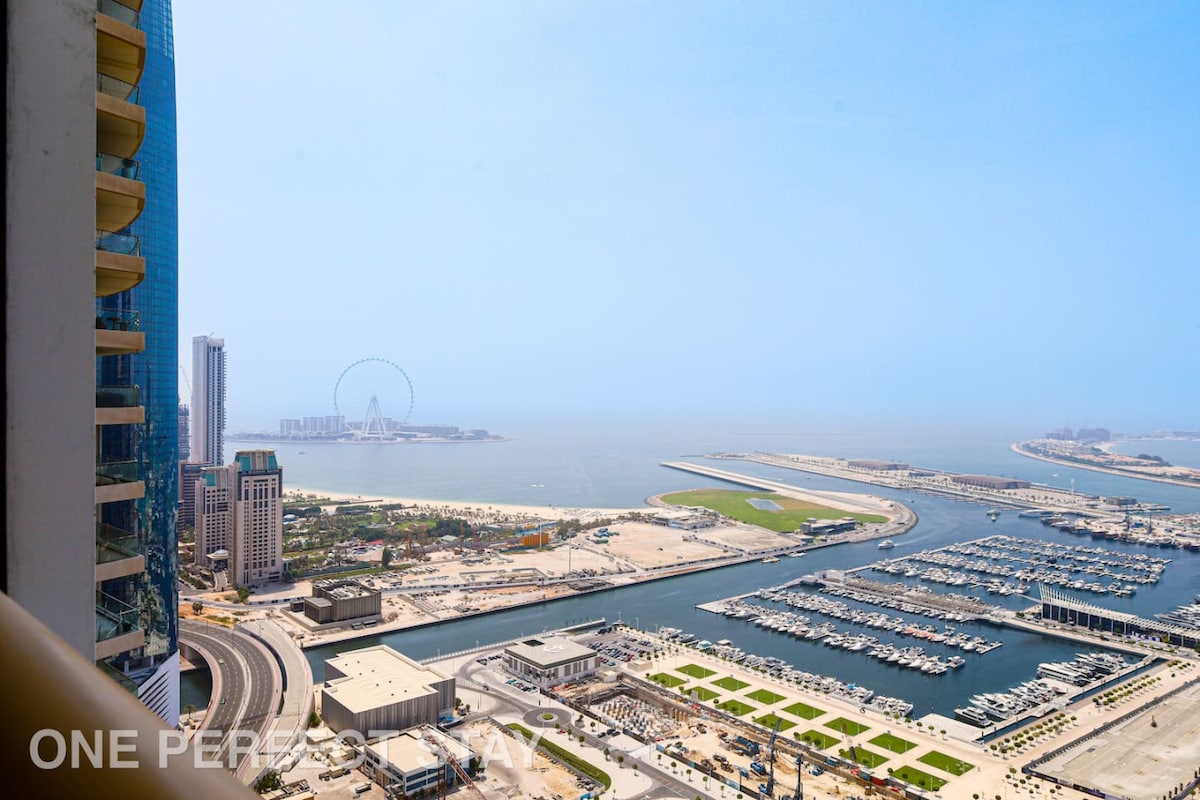 A panoramic view of Dubai Marina and the Arabian Gulf is showcased, featuring the iconic Ferris wheel in the distance. The image captures sprawling marina docks and sandy beaches, with clear blue skies enhancing the vibrant coastal scenery.
