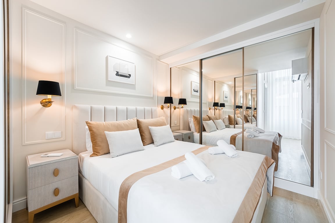 A well-appointed bedroom features a spacious queen bed with neutral-colored linens and decorative pillows. Mirrored wardrobes reflect the room's design, enhancing its brightness. A bedside table is positioned next to the bed, illuminating the space with twin wall sconces.