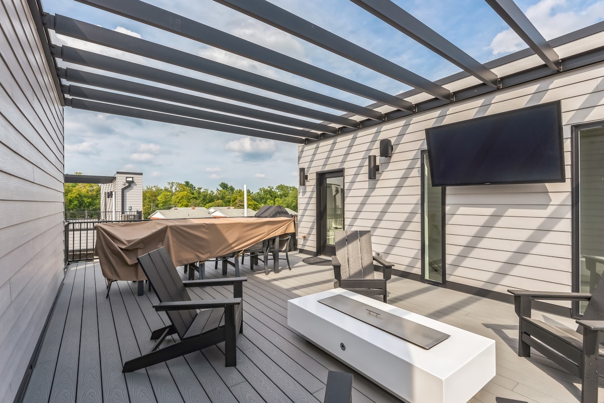 A spacious rooftop patio is highlighted, featuring modern outdoor furniture, including several chairs and a large dining table. A covered area with a grill is visible. Above, a retractable awning provides shade, while a wall-mounted TV is positioned for viewing.
