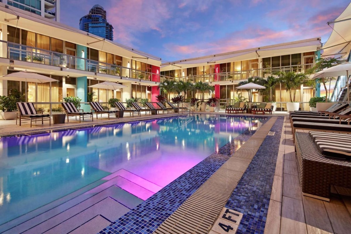 The outdoor pool area features modern lounge chairs arranged around the water, surrounded by palm trees and colorful illuminated buildings. Umbrellas provide shade, while gentle ripples on the pool's surface reflect the evening sky's pastel hues.