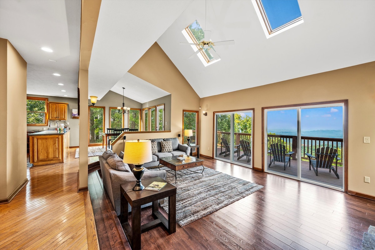 An open-concept living area features large windows offering mountain views and abundant natural light. A comfortable seating arrangement is complemented by a coffee table and a warm area rug. Skylights enhance the space, while a dining area and a view of the kitchen are visible in the background.