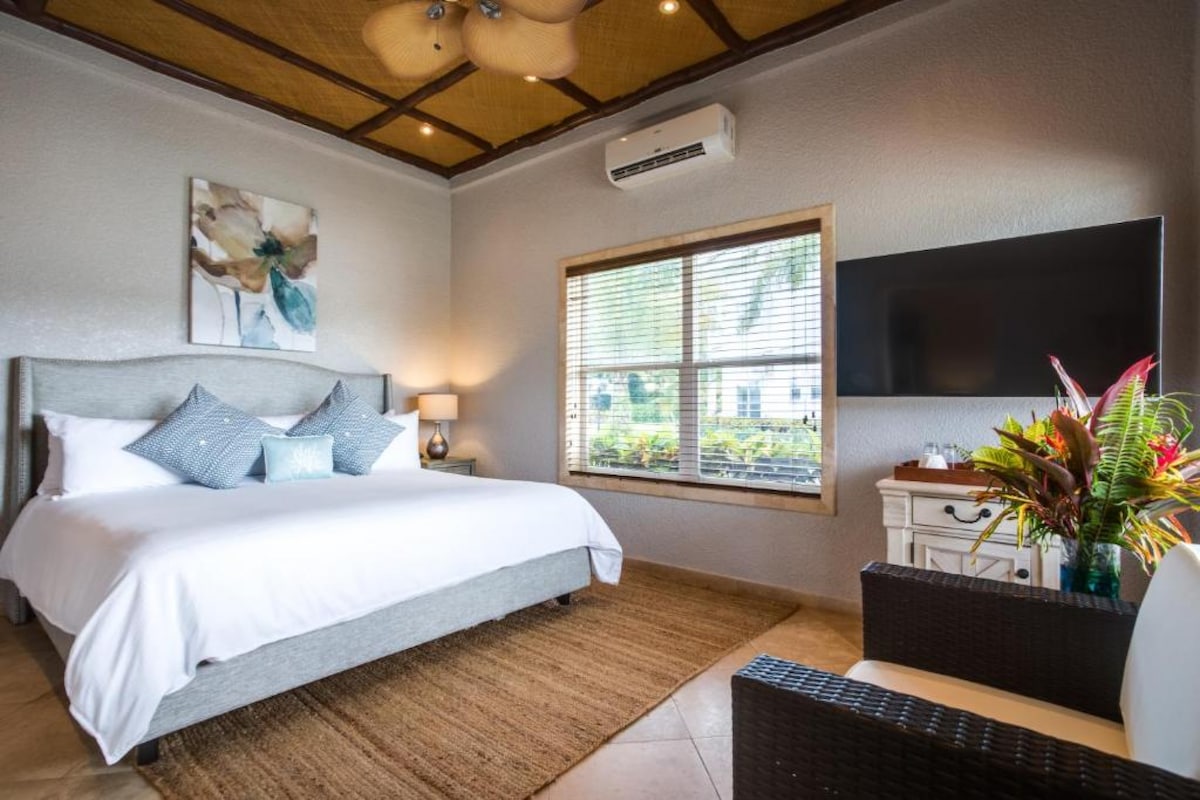 A spacious bedroom features a large bed with crisp white linens and decorative pillows. Natural light floods the room through a window adorned with horizontal blinds. A comfortable seating area and a modern entertainment console are also visible, complemented by tropical plants.