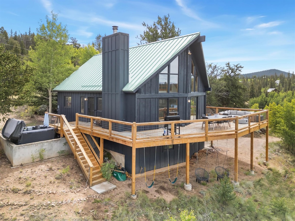 An exterior view of a modern cabin showcases a metal roof and large glass windows that offer scenic mountain views. A spacious wooden deck features seating areas and a hot tub, with a staircase leading down to a yard featuring swings and surrounding greenery.
