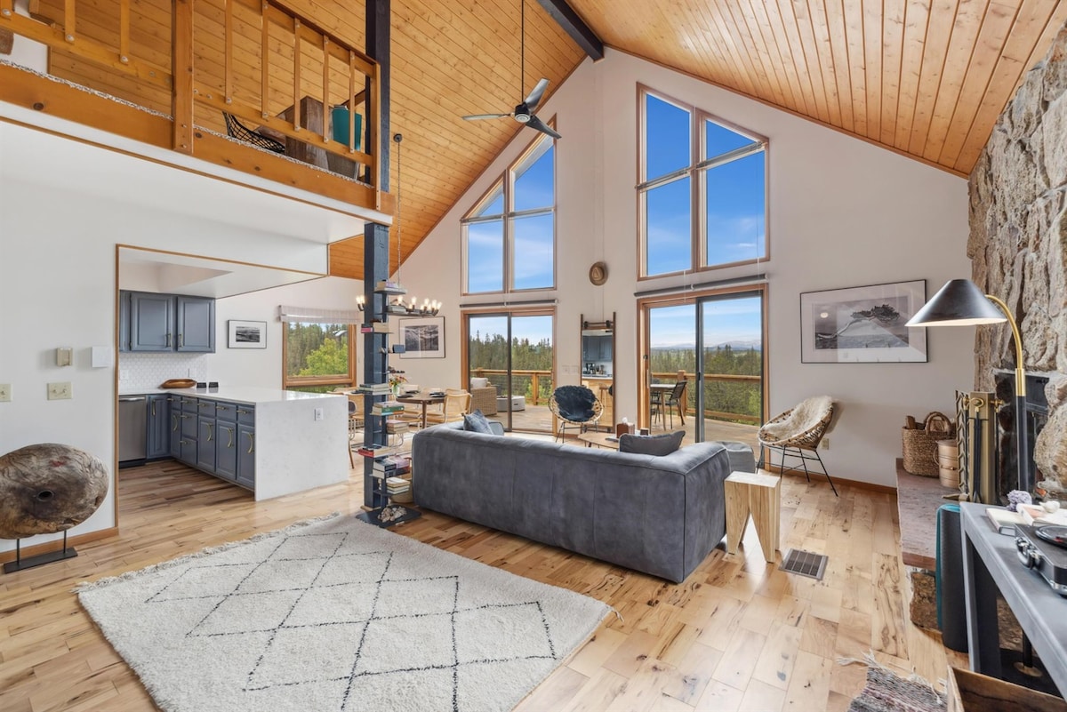 A spacious living area is illuminated by large windows, providing a view of the surrounding nature. The interior features wooden floors, a cozy seating arrangement, and a stone fireplace. A dining area is visible in the background, enhancing the inviting atmosphere.