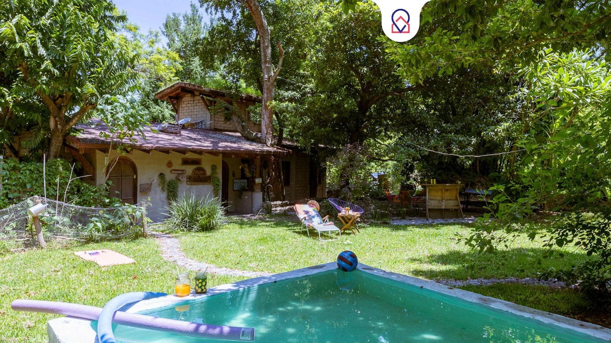 A refreshing swimming pool is set in a lush garden, surrounded by greenery. The house features a rustic design with a sloping roof and wooden elements visible from the outdoor area. Comfortable loungers are positioned on the grass, inviting relaxation.