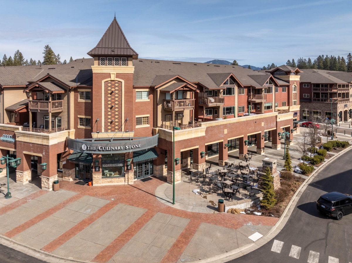 The image captures a multi-story brick building with a café called The Culinary Stone prominently displayed on the ground level. Outdoor seating areas are visible along the sidewalk, with a mix of tables and chairs, creating a vibrant atmosphere. Surrounding trees and mountain views enhance the setting.