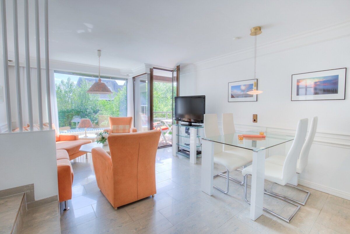 The bright living and dining area features a glass table surrounded by white chairs, with an orange sofa set positioned nearby. Large windows offer access to a lush outdoor view, while a wall-mounted TV is visible for entertainment.