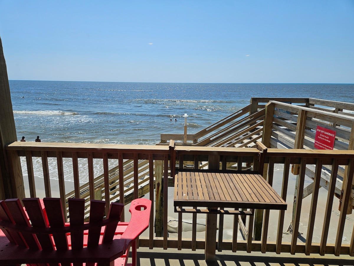 A private balcony offers a clear view of the ocean, with wooden railings framing the scene. A table is placed beside a pair of red chairs, creating a relaxed setting. Gentle waves and a sandy beach are visible, with guests enjoying the water in the distance.