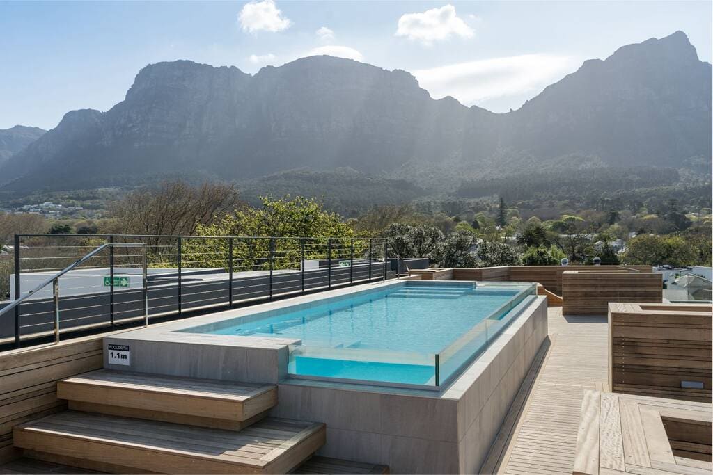A rooftop pool is displayed against a backdrop of striking mountains, with wooden decking surrounding its perimeter. Lush greenery can be seen in the distance, and the sky is bright with scattered clouds.