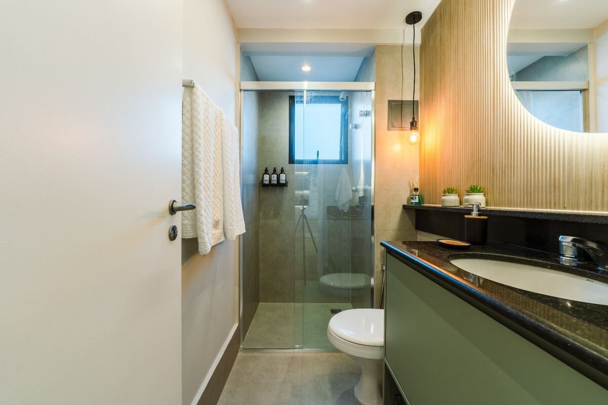 A modern bathroom features a glass shower enclosure and a sleek toilet. Soft white towels are hung on the wall, while a round mirror above the basin adds depth. Minimalist decor is complemented by subtle greenery on the countertop.