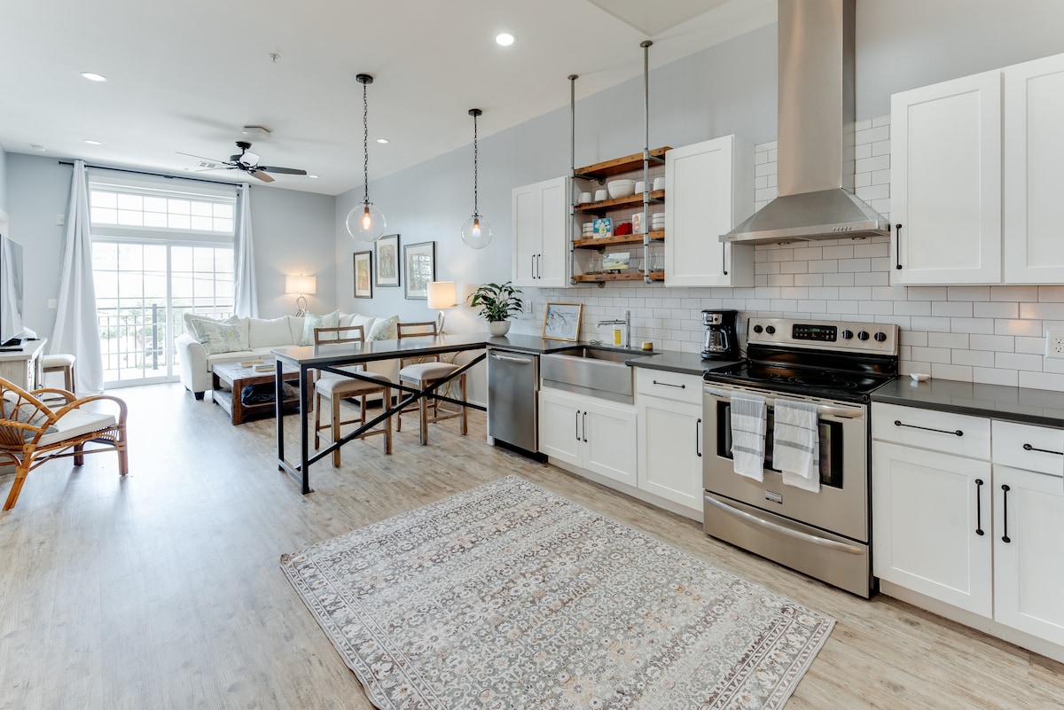 The modern kitchen features stainless steel appliances, including an oven and refrigerator, set against white cabinetry. A dining table is positioned near the living area, with comfortable seating visible in the background. Large windows allow natural light to brighten the space, complemented by stylish pendant lighting.