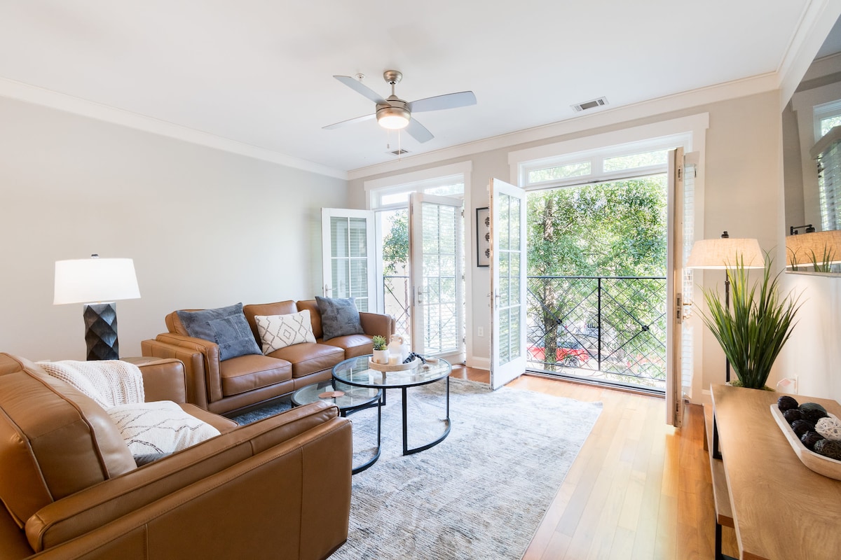 A light-filled living area features two comfortable leather sofas and a round glass coffee table. French doors open to a balcony overlooking greenery, while a ceiling fan provides a gentle breeze. A floor lamp and decorative plants enhance the welcoming space.