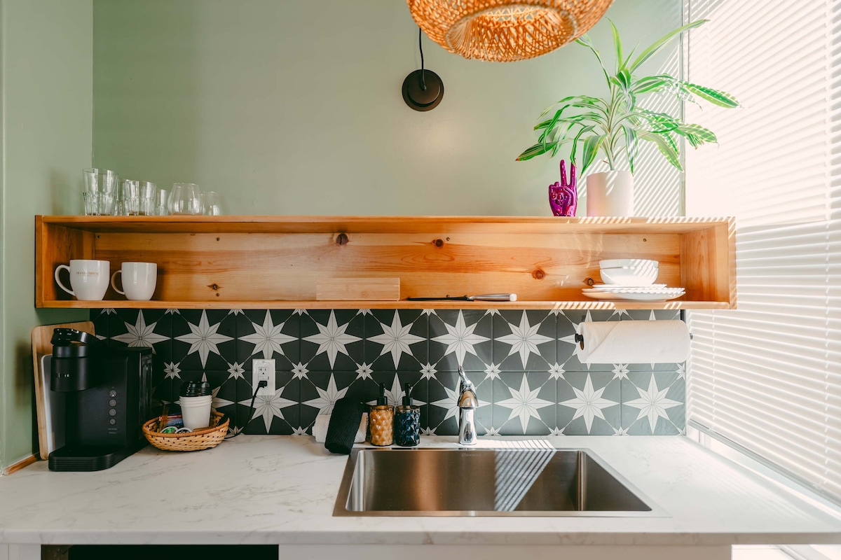 A kitchenette showcases a sleek sink beneath a wooden shelf displaying various dishware. A coffee maker and a decorative basket sit on the countertop, with a striking patterned backsplash enhancing the space. Natural light filters through the window, complemented by green plants.