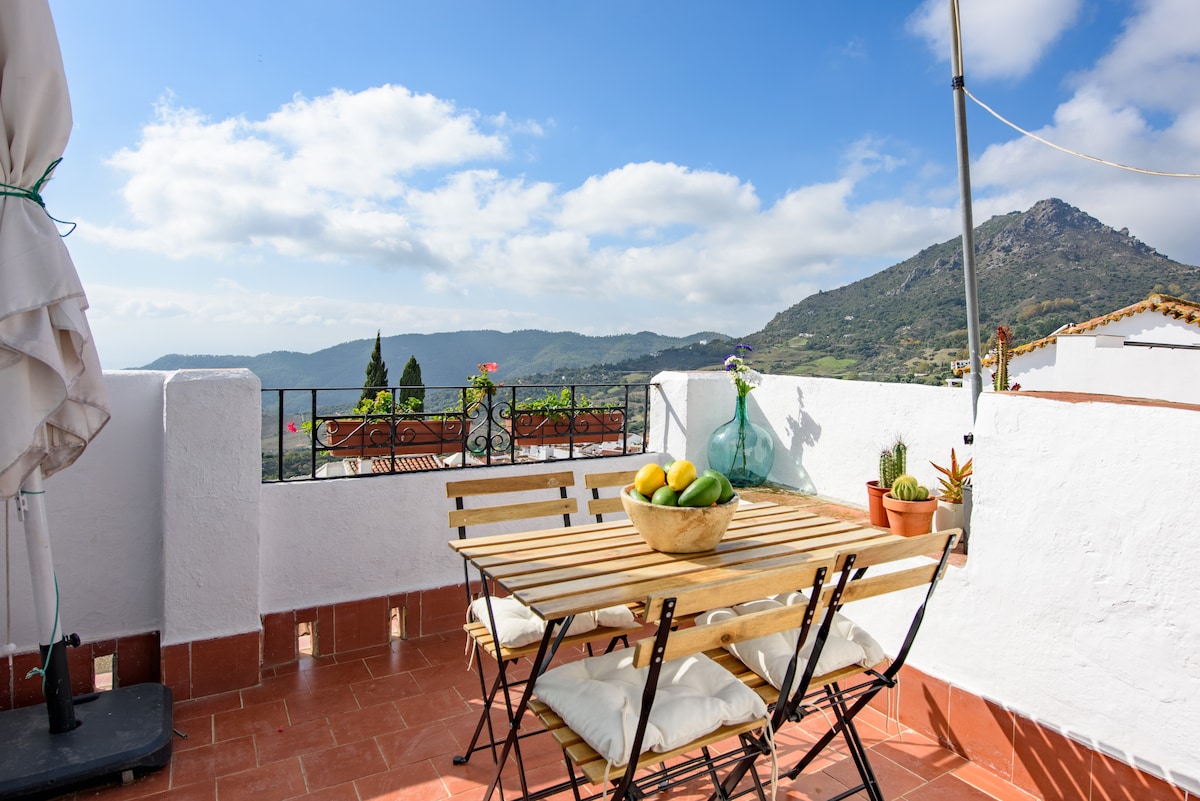 An outdoor terrace is shown with a wooden table surrounded by four folding chairs. A bowl of fruit is centered on the table. Scenic mountain views and a partly cloudy sky create a serene backdrop. Potted plants add a touch of greenery to the space.
