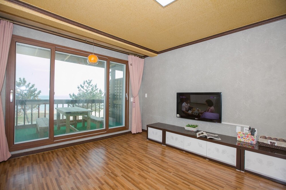 A spacious living area is depicted, featuring large sliding glass doors that allow natural light to fill the space. A modern television is mounted on a low cabinet, with minimal decor on the surface. Light wooden flooring complements the neutral-colored walls.
