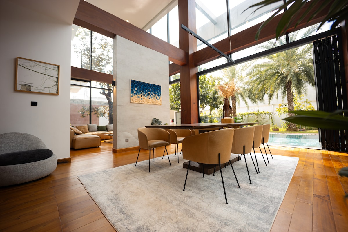 The dining area is characterized by a large wooden table surrounded by six tan chairs. Natural light floods the space through expansive glass walls, offering views of the landscaped garden and private pool. A soft area rug complements the warm wooden flooring.