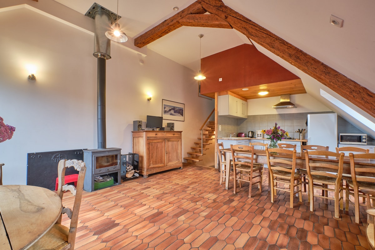 An inviting open-plan living area features warm terracotta flooring and exposed wooden beams. A wooden dining table with multiple chairs is positioned near a well-equipped kitchen. A cozy stove stands in one corner, complementing the overall rustic charm of the space.