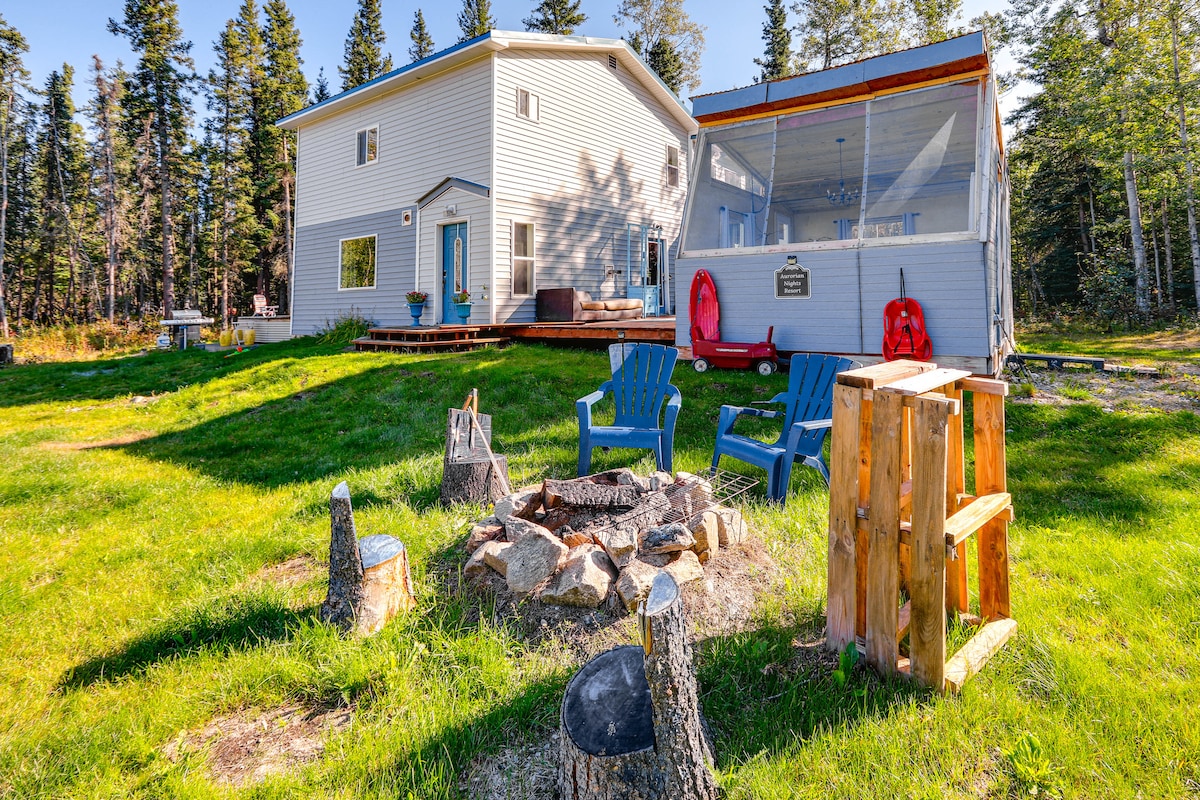A grassy area features a fire pit surrounded by log seats and wooden stools. The two-story vacation rental is visible in the background, alongside a screened outdoor sitting area. Tall trees provide a natural backdrop to the spacious yard.