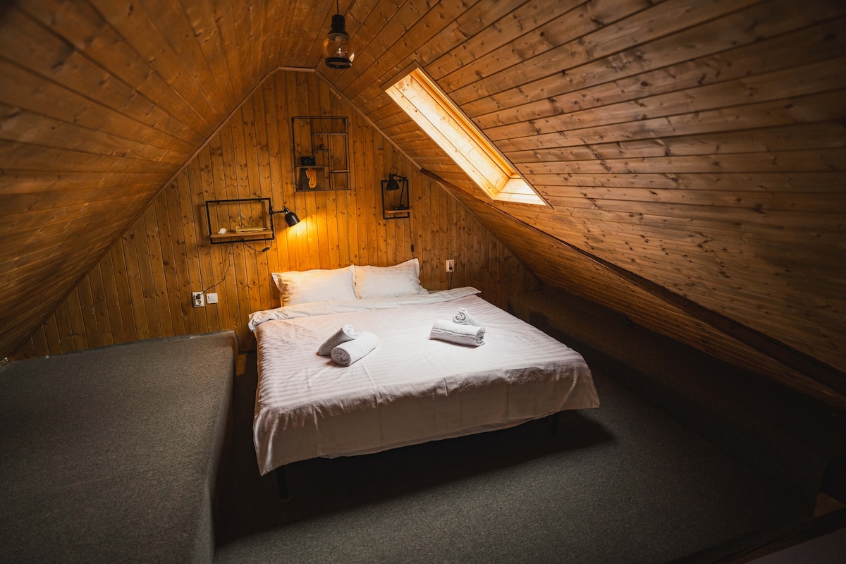 An attic bedroom features a spacious bed with neatly arranged towels. The wooden walls and sloped ceiling create a cozy atmosphere, while natural light pours in from a skylight. A soft carpet adds comfort to the room.