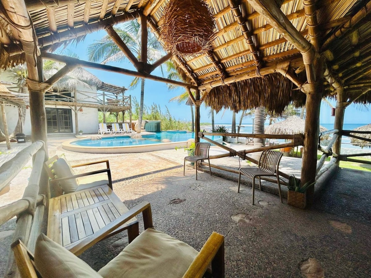 A spacious outdoor seating area is shaded by a thatched roof, featuring wooden furniture arranged for relaxation. A pool can be seen in the background, surrounded by lounges, with palm trees lining the edge. Natural light illuminates the cozy gathering space.