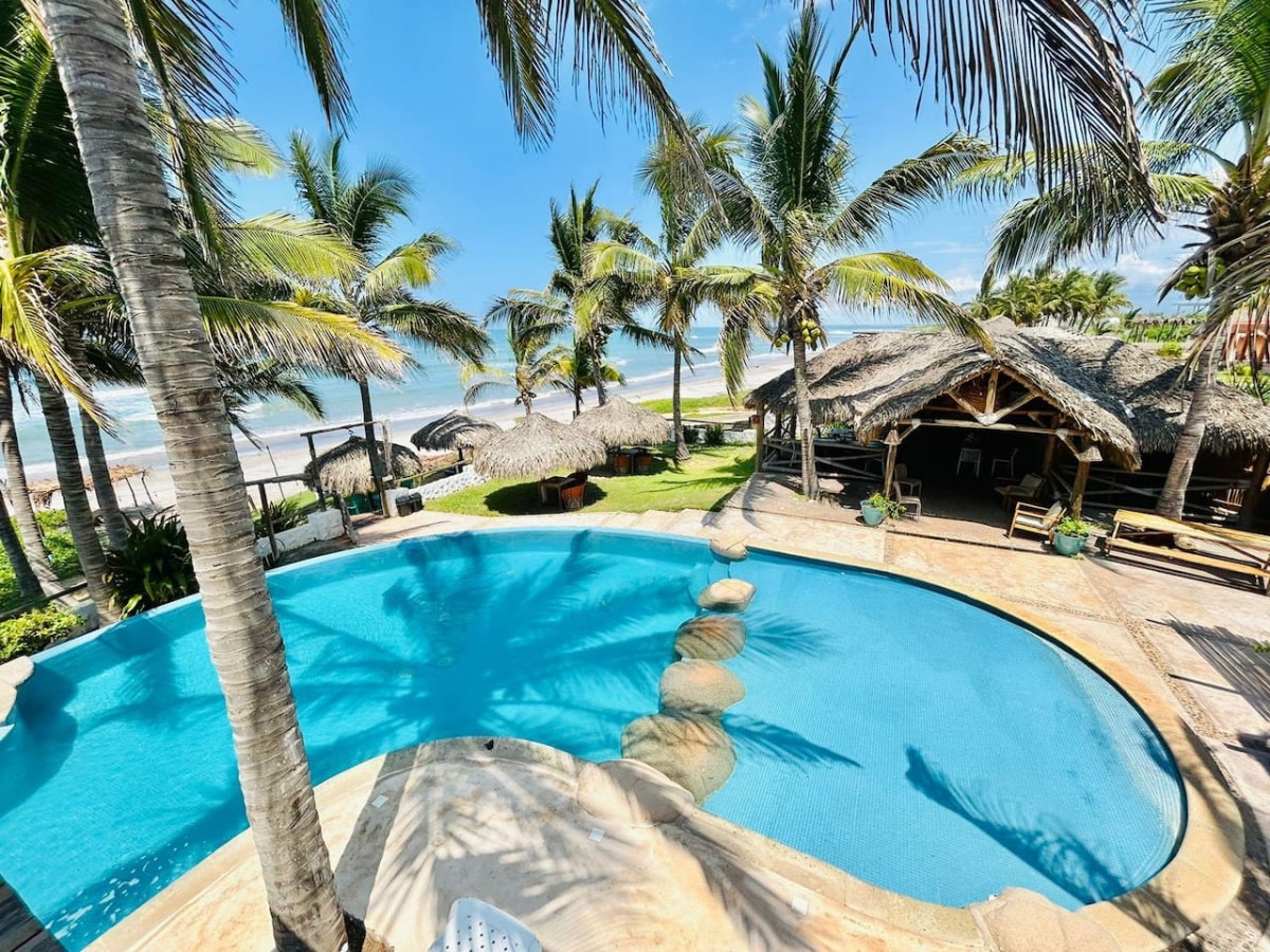 A large swimming pool is framed by palm trees and lounge chairs, with a thatched-roof structure visible nearby. The pool's clear water reflects the sky, while the beach and ocean are seen in the background, creating a serene atmosphere.