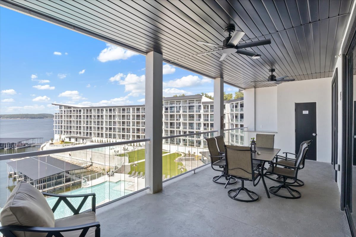 A spacious balcony features a dining area with a table and six chairs, overlooking the lake and community pool. Ceiling fans offer comfort, and the expansive views of the water and surrounding greenery can be appreciated from this outdoor space.