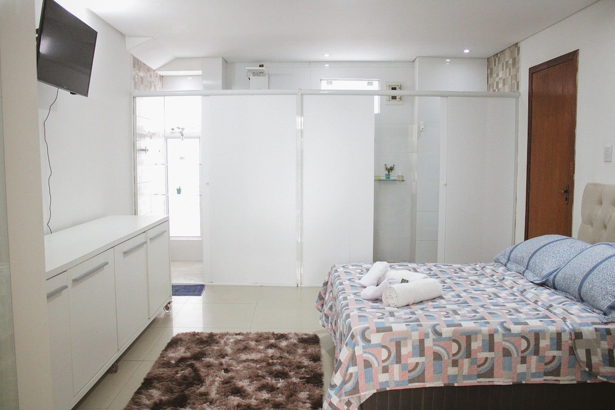 A bedroom is presented with a spacious bed featuring a patterned blanket. A television is mounted on the wall opposite the bed. The space includes white screens dividing the area from a modern bathroom, with a cozy rug on the floor adding warmth.