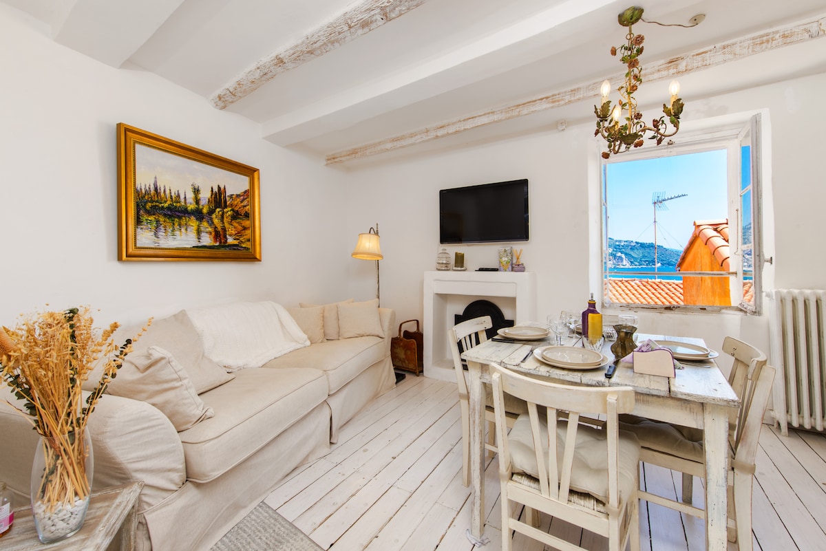A bright living area features a comfortable sofa and a dining table set for two. Natural light enters through a window, providing a view of the surrounding rooftops and hills. A television hangs on the wall, and decorative elements create a cozy ambiance.