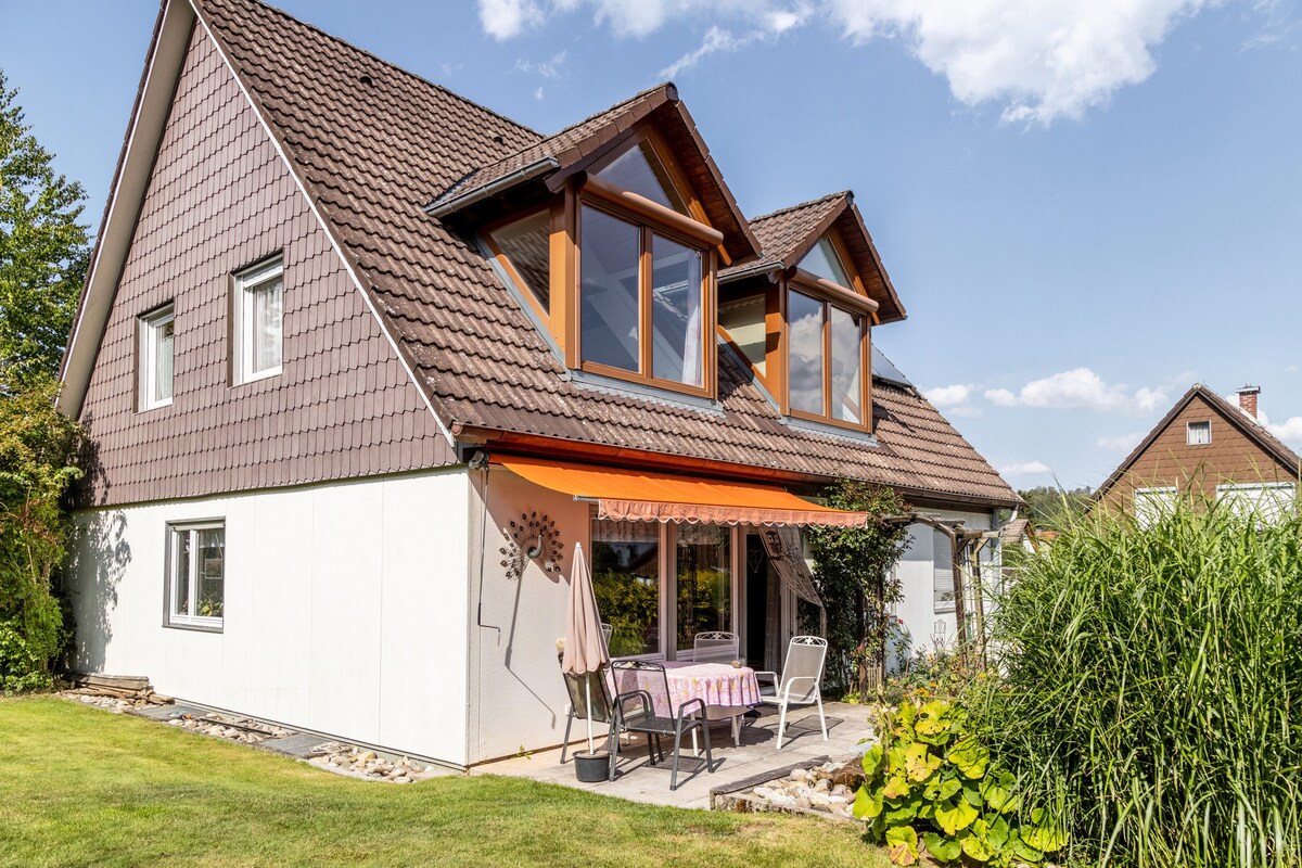 The exterior of the holiday apartment is showcased, featuring a two-story building with a combination of brown shingle roofing and light-colored walls. A shaded patio area is visible, furnished with a small table and chairs, surrounded by well-maintained greenery.