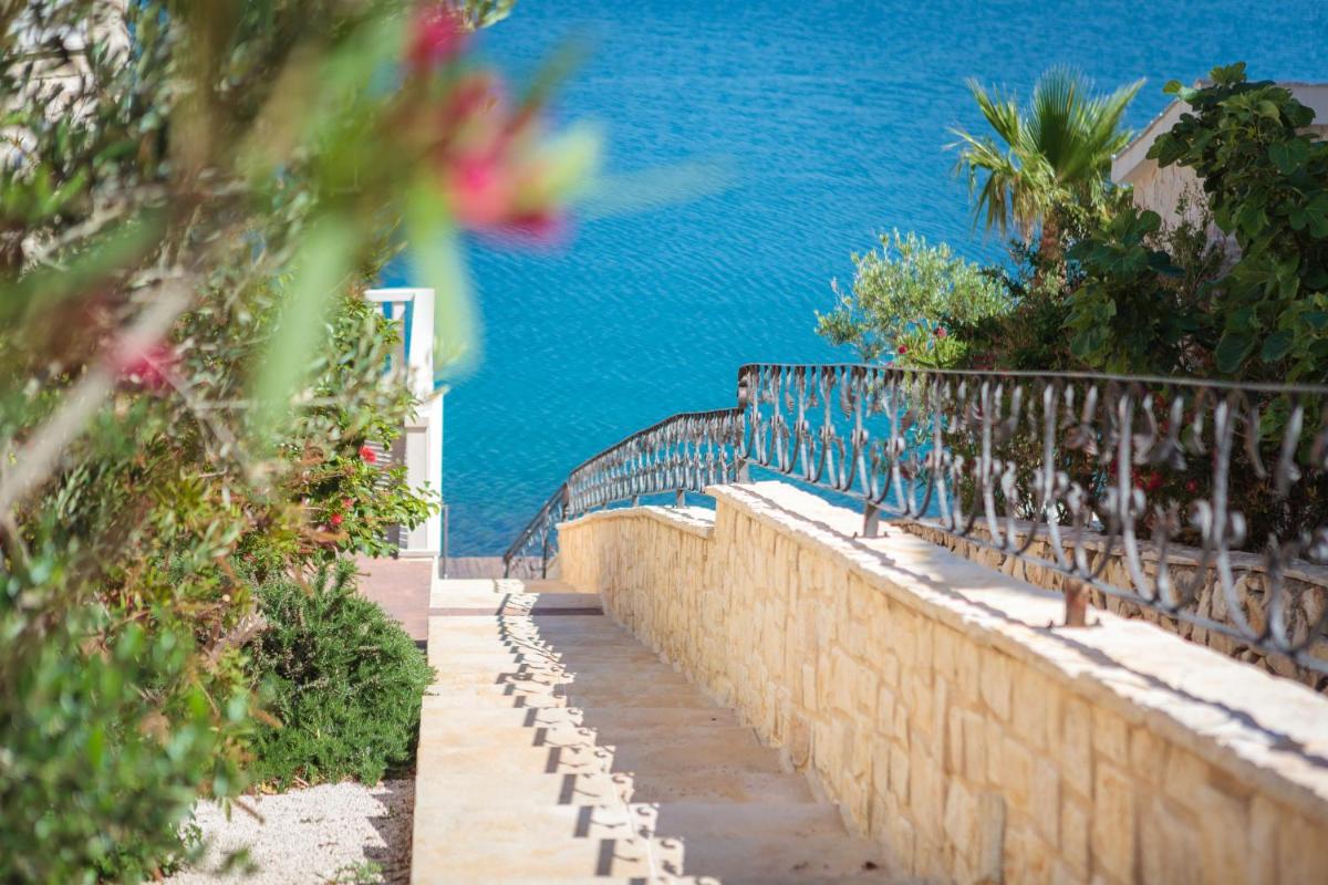 A gently curved stone pathway descends towards a vibrant blue body of water. Lush greenery and flowering plants border the path, complemented by a decorative iron railing. The scene captures a sense of tranquility and invites exploration toward the water's edge.