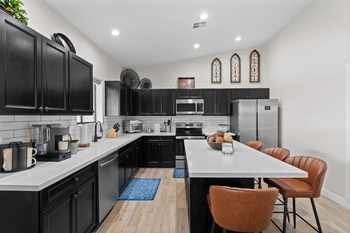 A modern kitchen is presented with dark cabinetry and a large central island. Stainless steel appliances, including a microwave and oven, are featured. Bar stools are lined up at the island, and decorative items are arranged along the countertops, creating a functional cooking space.